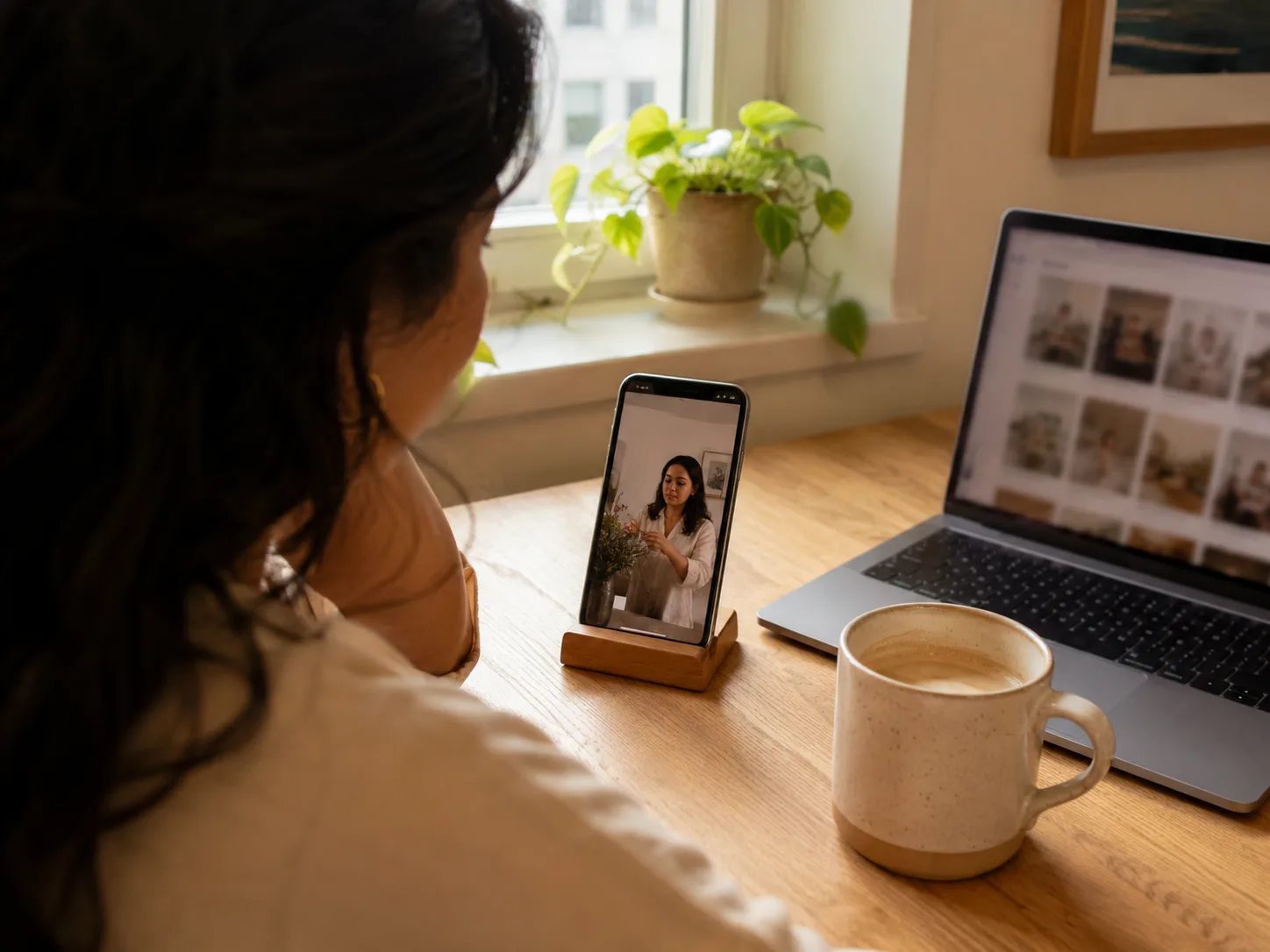 A creator watching a published short-form video play on a propped phone beside a laptop grid of posts, warm sunlit corner